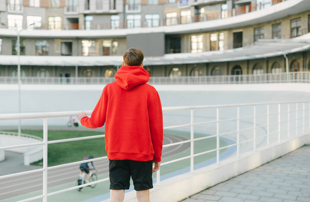 Back of a young man in a red hoodie stands on the street and looks at the velodrome. Man watches as bicycles ride on the velodrome, view from the back, background. - Photo, Image