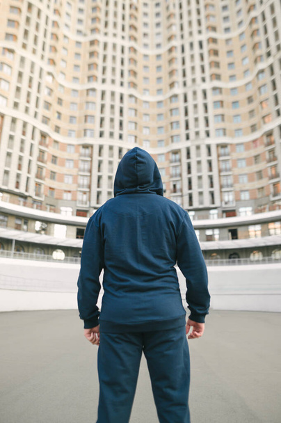 Mavi spor giyim ve kapüşonlu atletik adam mimarinin arka planında duruyor ve yukarı bakıyor, arkadan bakıyor. Dikey. - Fotoğraf, Görsel