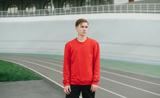 Portrait of a handsome young man in a red sweatshirt stands on the background of sports tracks and resting after a workout, looking away with a serious face. - 写真・画像