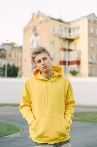 Portrait of stylish handsome young man in yellow hoodie posing at camera on cityscape background. Trendy street guy in casual clothes posing at camera. - Fotó, kép