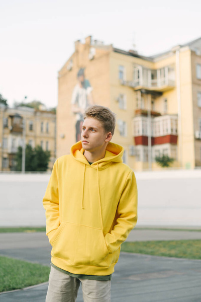 Portrait of stylish young man in yellow hoodie posing at camera on background of velodrome and old buildings. Vertical. - 写真・画像