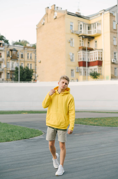 Egy fiatalemberről készült teljes hosszúságú fotó stílusos nyári ruhákban, a játszótéren sétálva a régi város építészetének hátterében, félrenézve. Függőleges - Fotó, kép