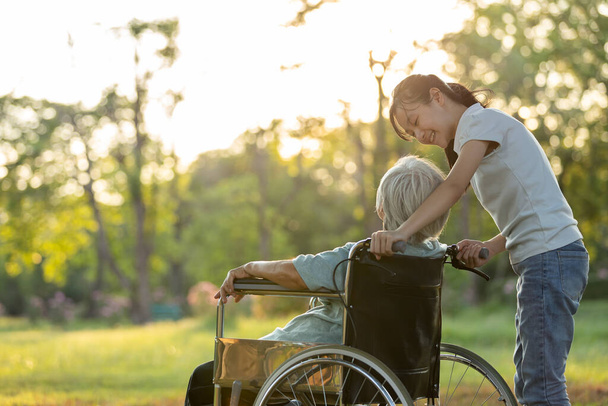 Ευτυχισμένη ηλικιωμένη ηλικιωμένη γυναίκα σε αναπηρικό καροτσάκι με την εγγονή της να περπατάει στο πάρκο, χαμογελώντας κοριτσάκι και ηλικιωμένη γιαγιά να ξεκουράζονται και να μιλάνε μαζί το βράδυ στο ηλιοβασίλεμα φύση, αγάπη, οικογενειακές σχέσεις - Φωτογραφία, εικόνα