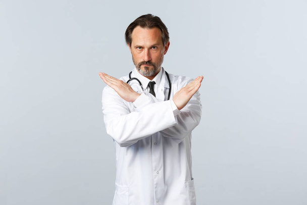 Covid-19, coronavirus outbreak, healthcare workers and pandemic concept. Serious displeased male doctor in white coat, cross hands to show stop gesture, prohibit or forbid smth - Fotoğraf, Görsel