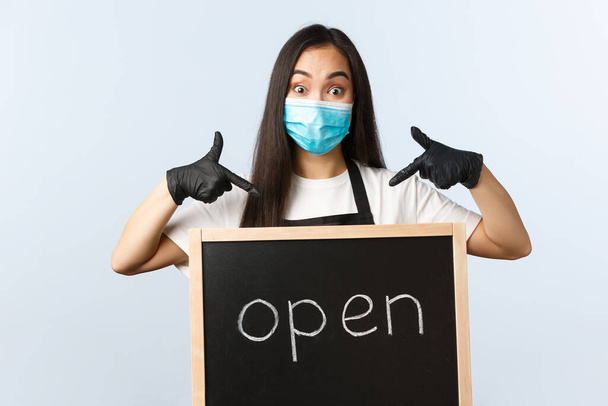 Small business, covid-19 pandemic, preventing virus and employees concept. Excited cute asian female barista, cafe or store worker in medical mask pointing at we are open sign - Foto, imagen