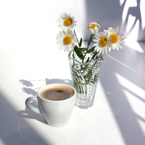 Ένα φλιτζάνι μαύρο καφέ με αφρό, ένα μπουκέτο λευκά χαμομήλια λουλούδια σε ένα κρυστάλλινο βάζο με νερό σε ένα λευκό τραπέζι στο φως του ήλιου και όμορφες σγουρές σκιές - Φωτογραφία, εικόνα