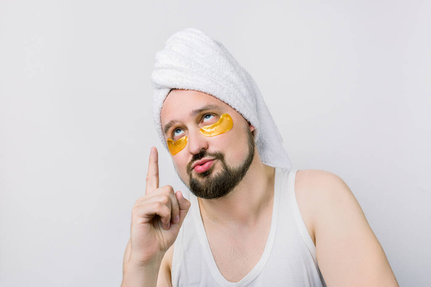 Beauty treatment, skin care, metrosexual concept. Handsome bearded man with golden eye patches under eyes and white towel on head, posing on white background with confused questionable expression - Photo, Image