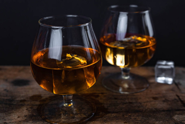 Two whiskey / cognac glasses with ice on a wooden background. Dark backdrop. - Photo, Image