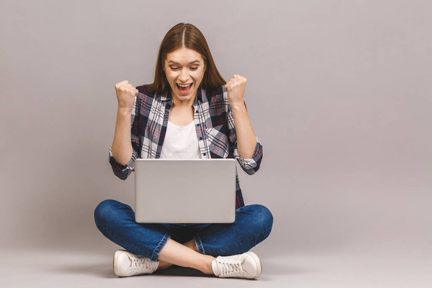 Vencedor! Garota sorridente animado sentado no chão com laptop, levantando uma mão no ar é que ela ganha, isolado em fundo cinza
. - Foto, Imagem