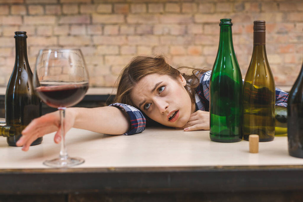 Mladá smutná a vyčerpaná alkoholička sedící v kuchyni na gauči a popíjející červené víno držící sklenici úplně opilá, vypadající sklíčeně a trpící kocovinou v alkoholismu a zneužívání alkoholu. - Fotografie, Obrázek