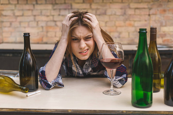 Junge traurige und vergeudete Alkoholikerin, die auf der Küchencouch sitzt und Rotwein trinkt, völlig betrunken und deprimiert wirkt und an Alkoholismus und Alkoholmissbrauch leidet. - Foto, Bild