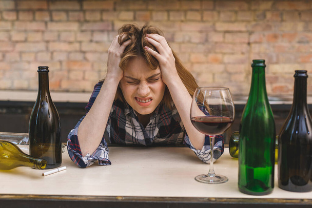 Νεαρή θλιμμένη και μεθυσμένη αλκοολική που κάθεται στον καναπέ της κουζίνας πίνοντας κόκκινο κρασί, εντελώς μεθυσμένη που δείχνει θλιμμένη μοναχική και υποφέρει από πονοκέφαλο από αλκοολισμό και κατάχρηση αλκοόλ. - Φωτογραφία, εικόνα