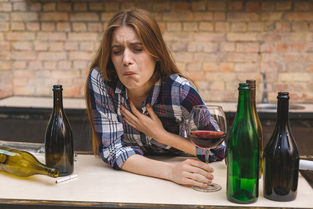 Jeune femme alcoolique triste et gaspillée assise sur le canapé de la cuisine à boire du vin rouge, complètement ivre regardant déprimé solitaire et souffrant de gueule de bois dans l'alcoolisme et l'abus d'alcool. - Photo, image