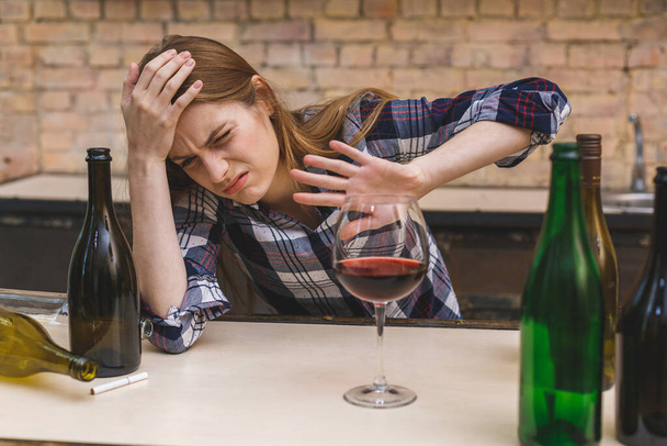 Молодая грустная алкоголичка, сидящая на кухонном диване, пьющая красное вино, совершенно пьяная, выглядящая подавленной одинокой и страдающей похмельем от алкоголизма и злоупотребления алкоголем
. - Фото, изображение