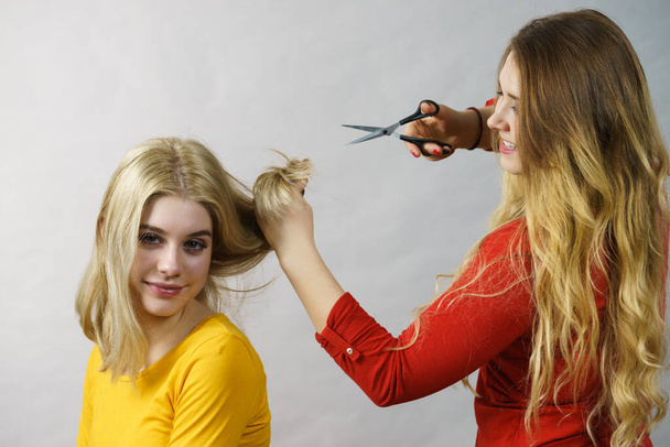Hairstyle and haircut. Young female barber holding scissors tool ready to trimming hair her friends. Two girls creating new hairdo coiffure - Foto, Imagem