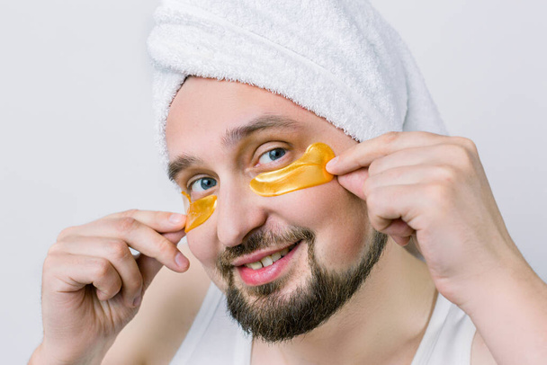 Smiling bearded man with white towel on head and with golden patches under his eyes, caring for his facial skin looking at camera on white background. Mens facial Spa. Concept of cosmetology for men. - Фото, изображение