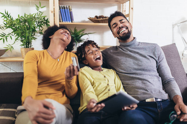 Linda niña afroamericana y sus hermosos padres jóvenes utilizando la tableta digital y haciendo compras en línea mientras se sienta en un sofá en casa
. - Foto, imagen