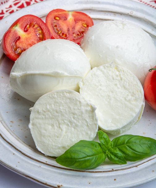 Mozzarella italienne au fromage à pâte molle, fromage blanc à base de lait de vache ou de buffle avec de l'herbe de basilic vert frais et de la tomate rouge en gros plan - Photo, image
