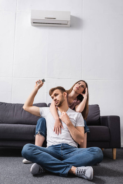 casal confuso com controlador remoto sofrendo de calor enquanto sentado em casa com ar condicionado quebrado
  - Foto, Imagem