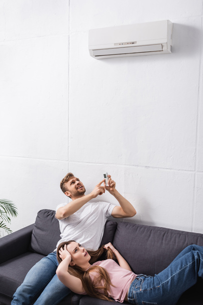 worried couple with remote controller suffering from heat at home with broken air conditioner  - Фото, изображение