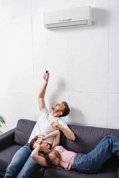 frustrated young couple with remote controller suffering from heat at home with broken air conditioner  - Foto, Bild