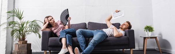 young couple with hand fan and newspaper suffering from heat at home, panoramic orientation - Fotoğraf, Görsel