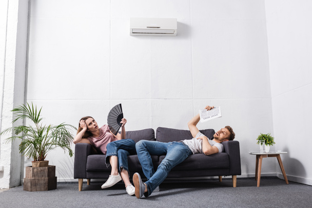 mladý pár s ruční ventilátor a noviny trpí horkem doma s rozbitou klimatizací  - Fotografie, Obrázek