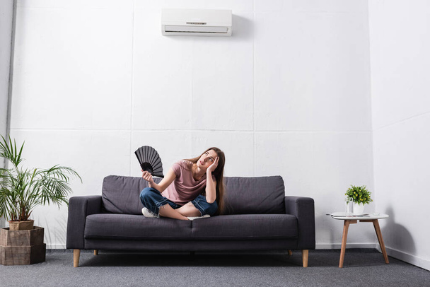 verwarde jonge vrouw die last heeft van warmte en het gebruik van de hand ventilator thuis met gebroken conditioner - Foto, afbeelding