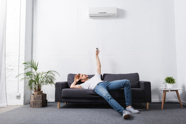 exhausted man holding remote controller and suffering from heat with broken air conditioner at home  - Fotografie, Obrázek