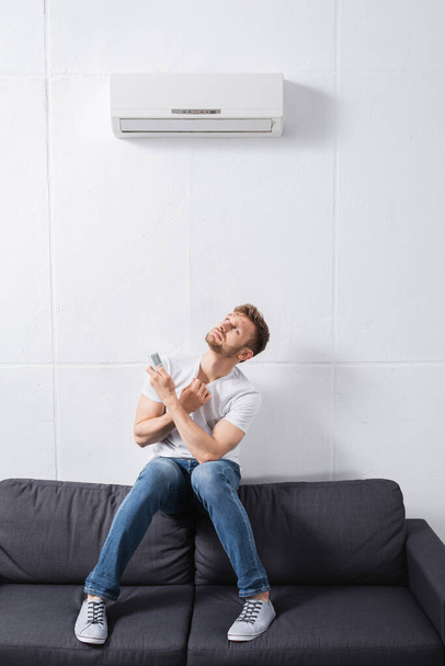homme confus tenant télécommande et souffrant de chaleur avec climatiseur cassé à la maison  - Photo, image