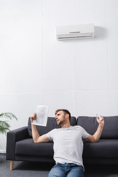 sad young man suffering from heat while using newspaper as hand fan at home with broken air conditioner - Фото, изображение