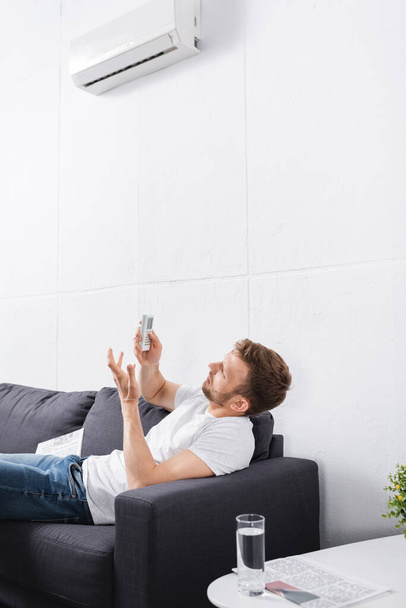 confused man trying to switch on air conditioner with remote controller at home  - Foto, Bild