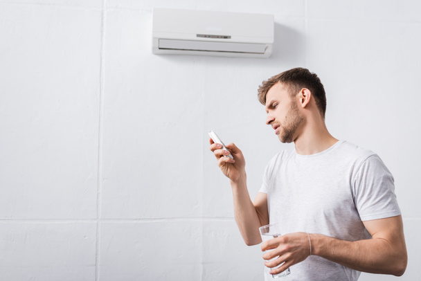 frustrado hombre sosteniendo un vaso de agua mientras intenta encender el aire acondicionado con el mando a distancia roto en casa
  - Foto, imagen