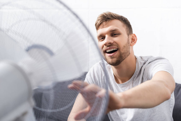przystojny pozytywny mężczyzna czuje się komfortowo z elektryczny wentylator w domu podczas letni ciepło  - Zdjęcie, obraz
