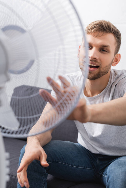 красивий щасливий чоловік відчуває себе комфортно з електричним вентилятором вдома під час літньої спеки
  - Фото, зображення