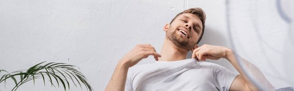 pozitivní emocionální muž se cítí pohodlně s elektrickým ventilátorem doma během letního vedra, panoramatické plodiny - Fotografie, Obrázek