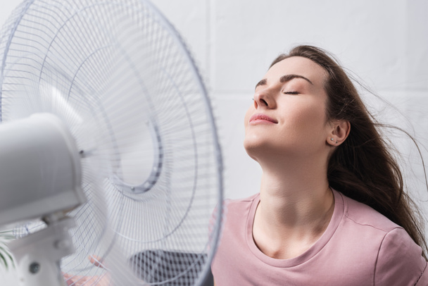 приваблива позитивна дівчина відчуває себе комфортно з електричним вентилятором вдома під час літньої спеки
  - Фото, зображення