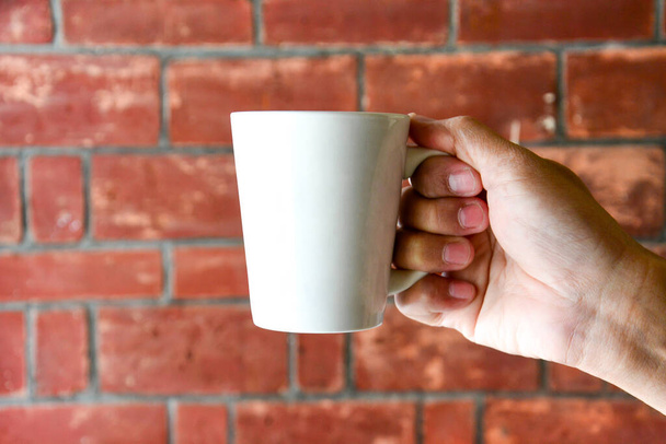 close up of male hand holding blank white coffee or tea cup on old red brick wall background texture with morning light, copy space - Photo, Image