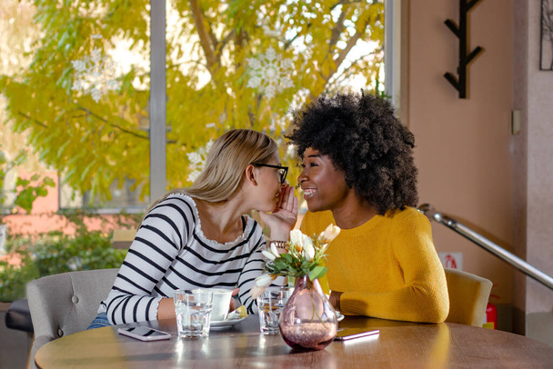 Dos amigas jóvenes disfrutando del café juntas en una cafetería, sentadas en una mesa, charlando y chismeando. Diversidad y concepto multiétnico
. - Foto, imagen