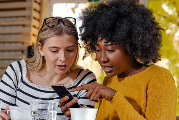 Dos amigas jóvenes disfrutando del café juntas en una cafetería, sentadas en una mesa, charlando y chismeando mientras rastrean las redes sociales en un teléfono inteligente. Diversidad y concepto multiétnico
. - Foto, Imagen
