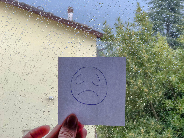 Hand holding a card with sad emoji written on it on the rainy window background - Photo, Image