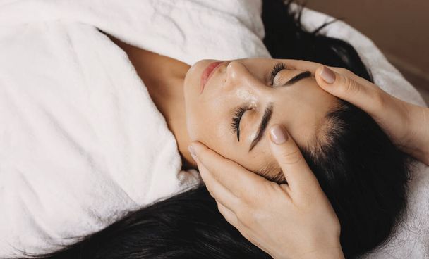 Mujer caucásica acostada en un centro de spa y teniendo una sesión de masaje en la cabeza con los ojos cerrados
 - Foto, Imagen