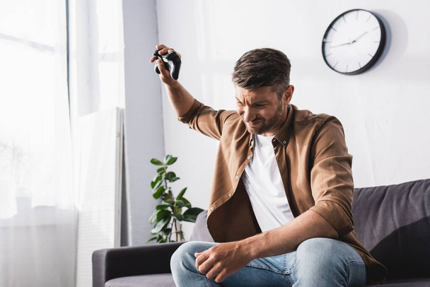 KYIV, UKRAINE - JUNE 9, 2020: upset, angry man throwing joystick while sitting on sofa at home - Foto, afbeelding