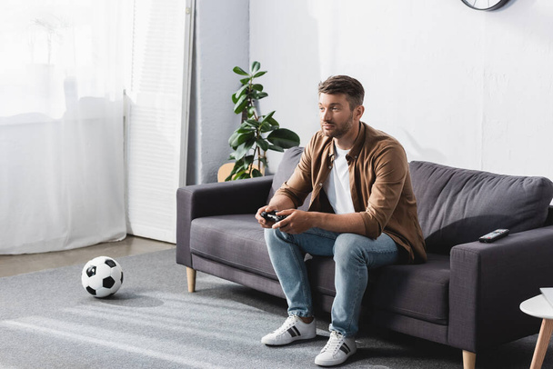 КИЕВ, УКРАИНА - 9 ИЮНЯ 2020: внимательный человек, играющий в видеоигру, сидя на диване возле футбольного мяча
 - Фото, изображение
