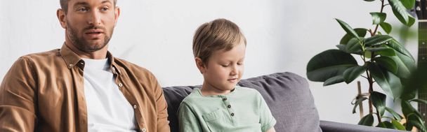 panoramic crop of thoughtful father and son sitting on sofa near potted plant - Photo, Image