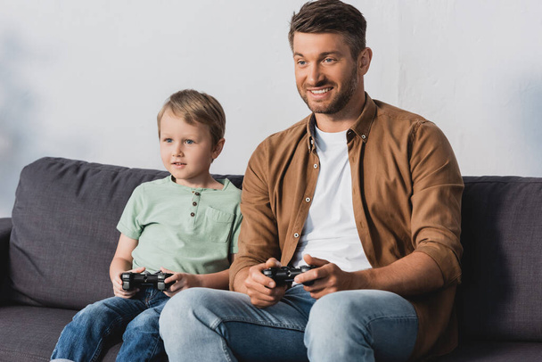 KYIV, UKRAINE - JUNE 9, 2020: smiling father and son sitting on sofa and playing video game with joystcks - Photo, Image