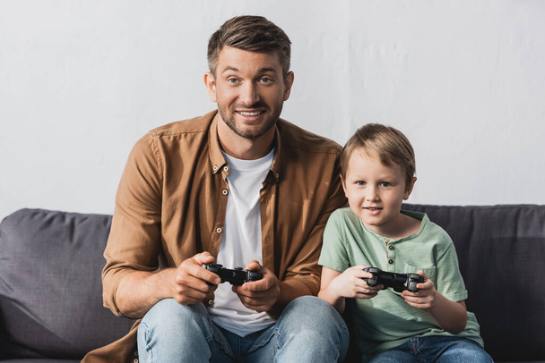 KYIV, UKRAINE - JUNE 9, 2020: excited father and son sitting on sofa and playing video game with joysticks - Photo, Image