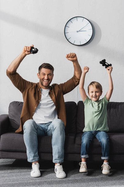 KYIV, UCRAINA - 9 GIUGNO 2020: padre e figlio eccitati che guardano la macchina fotografica tenendo i joystick e mostrando i gesti dei vincitori  - Foto, immagini