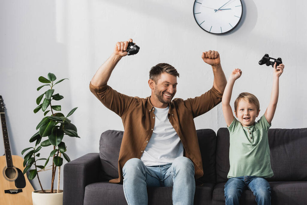 KYIV, UKRAINE - JUNE 9, 2020: happy father and son showing winner gestures while holding joysticks - Foto, imagen
