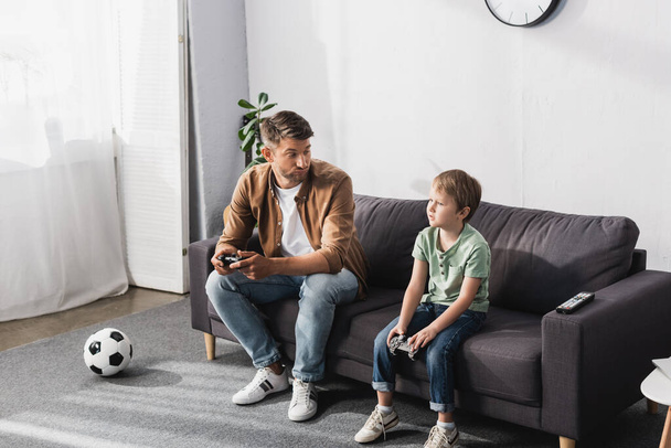 KYIV, UKRAINE - JUNE 9, 2020: displeased father looking at upset son while sitting on sofa with joysticks - Foto, imagen